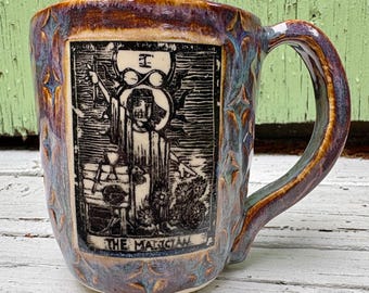 May include: A ceramic mug with a dark purple and blue glaze. The mug features a black and white illustration of The Magician tarot card. The handle and rim have a brown glaze. The mug is on a white surface.
