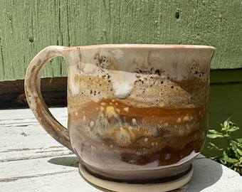 May include: A ceramic mug with a wide handle and a glossy finish. The exterior features a layered glaze pattern in shades of cream, tan, ochre, and dark brown, with speckled textures near the rim and a smooth, light-colored base at the bottom.