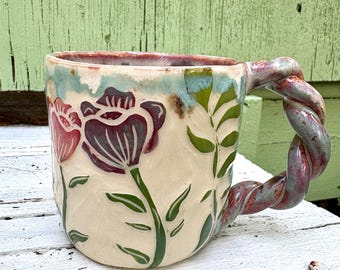 May include: A ceramic mug with a cream-colored base featuring hand-painted dark purple and red flowers with green stems and leaves. The mug has a distinctive twisted handle with a multi-colored glaze in shades of blue, purple, and red.