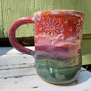 May include: A ceramic mug featuring a glossy glaze with a color gradient transitioning from orange at the top to purple in the middle and green at the base. The upper section is decorated with embossed flower patterns and small circular indentations.