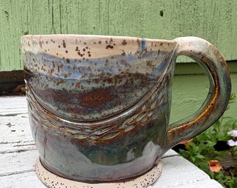 May include: A ceramic mug featuring a speckled cream-colored rim and base, with a central band of blended blue, green, and deep brown glaze. The body displays a textured, carved horizontal line pattern wrapping around the middle section.