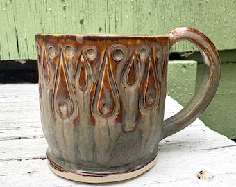 May include: A ceramic mug featuring a hand-thrown silhouette with a curved handle. The exterior displays a two-tone glaze with a warm brown upper section decorated with embossed teardrop shapes and a muted sage green base with vertical drips.