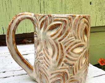 May include: A ceramic mug featuring a raised, carved floral pattern across its surface. The mug has a glossy, light beige glaze with darker brown accents highlighting the carved details and a curved handle on the left side of the vessel.