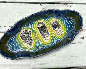 May include: An oval ceramic tray with a scalloped edge, featuring a deep blue and green glaze. The center displays three black ink-style illustrations of jellyfish surrounded by concentric yellow and white wavy lines on a blue background.