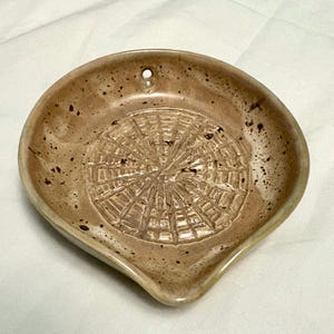 May include: A speckled, ceramic spoon rest in shades of tan and brown. The rest features a textured, circular design in the center and a small hole for hanging. The spoon rest has a curved shape with a small spout.