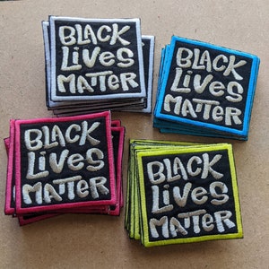 May include: A stack of four black fabric patches with white embroidered text that reads "Black Lives Matter". The patches are in different colors: white, blue, pink, and yellow.