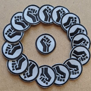 May include: A circle of 17 white and black iron-on patches with a raised fist design. The patches are arranged in a circle around a single patch in the center.