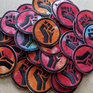 May include: A pile of colorful fabric patches with a raised black fist design. The patches are circular and have a tie-dye pattern.