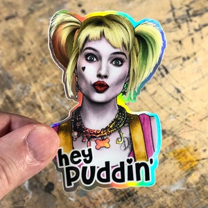 May include: A holographic sticker featuring Harley Quinn from the movie "Birds of Prey". The sticker has a rainbow iridescent background and features Harley Quinn with her signature pigtails and a pink and yellow outfit. The text "hey puddin'" is printed on the sticker.