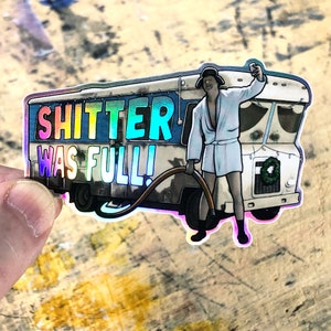 May include: A sticker featuring a cartoon illustration of a person in a white bathrobe standing next to a camper van. The text on the sticker reads "SHITTER WAS FULL!" in rainbow colors.