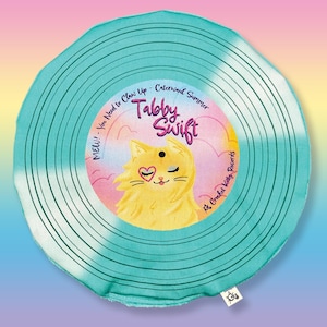 Könnte beinhalten: Ein rundes, türkisfarbenes Kissen, das einer Schallplatte nachempfunden ist. In der Mitte befindet sich eine Cartoon-Katze mit herzförmiger Brille und dem Text "Tabby Swift". Zusätzlicher Text lautet "MEOW! - You Need to Claw Up - Caterwaul Summer" und "Crochet Kitty Records."