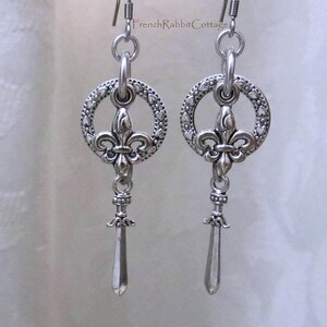 May include: Silver dangle earrings with a fleur-de-lis charm and a sword-shaped pendant. The charm is surrounded by a circle of small hearts.