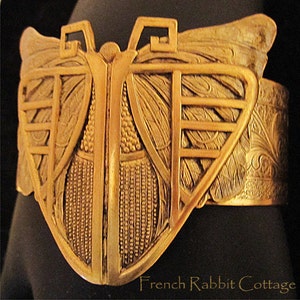 May include: A gold-toned metal butterfly bracelet with intricate details and a textured surface. The bracelet is open on one side and has a hinged clasp.