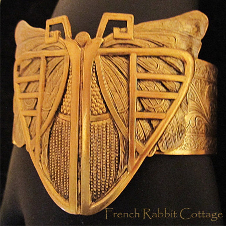 May include: A gold-toned metal butterfly bracelet with intricate details and a textured surface. The bracelet is open on one side and has a hinged clasp.
