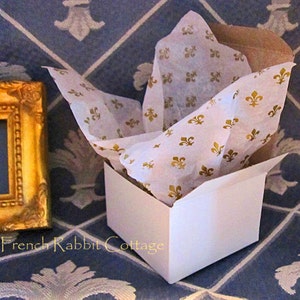 May include: A white gift box with a lid partially open, filled with white tissue paper printed with gold fleur-de-lis designs. The box is sitting on a blue and white patterned surface.