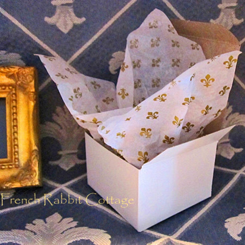 May include: A white gift box with a lid partially open, filled with white tissue paper printed with gold fleur-de-lis designs. The box is sitting on a blue and white patterned surface.