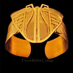 May include: A gold-toned metal ring with a butterfly design. The butterfly has intricate details and is centered on the ring. The ring is wide and has a smooth, polished finish.