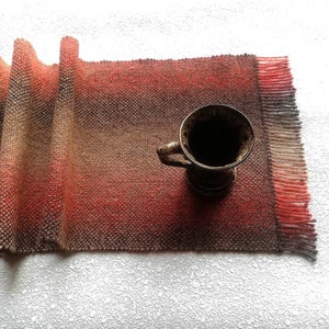 Könnte beinhalten: Ein gewebter Tischläufer mit einem Farbverlauf von Braun zu Rot und einem Fransenrand. Eine dunkelbraune Keramiktasse mit Henkel steht auf dem Läufer. Der Läufer besteht aus einem strukturierten Material.