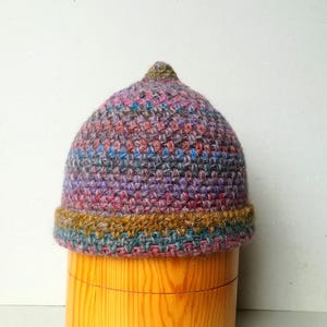 Puede incluir: Un gorro tejido a mano en forma de cúpula, con una mezcla de hilos morados, rosas, azules y grises. El borde es una banda de color marrón, verde azulado y dorado. El sombrero se exhibe en una forma de madera.