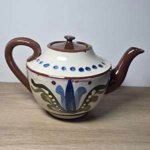 以下が含まれることがあります： クリーム色の陶器製ティーポット。茶色のハンドル、注ぎ口、蓋が付いています。ポットの前面には手描きの青と緑の花柄が施され、上部には青いドットの列が円状に並んでいます。