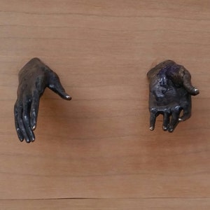 May include: Two bronze-colored hand sculptures mounted on a light brown wooden surface. One hand is open, palm down, and the other is in a fist with the index finger extended. The hands are detailed and appear to be cast.