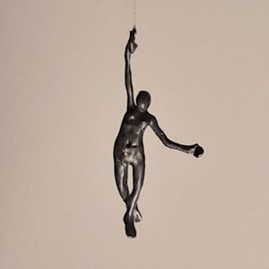 Könnte beinhalten: Eine schwarze Metallsculptur einer Person, die mit einem Arm nach oben und dem anderen ausgestreckt von der Decke hängt.