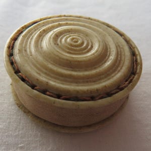Op de afbeelding: Een kleine, ronde, antieke sieradendoos. Het deksel heeft een spiraalvormig ontwerp. De doos is lichtbeige met een donkerbruine gestikte band. De doos staat op een witte ondergrond. Diameter ongeveer 5 cm.