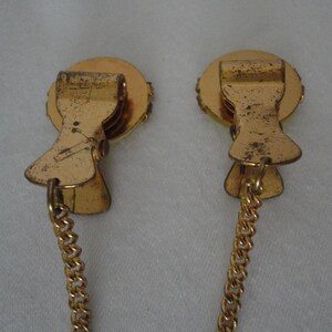 May include: A pair of gold-tone metal clips with a chain connecting them. Each clip has a round, decorative top with a raised design.