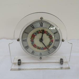 May include: A vintage clock with a floral design on the face, encased in a clear acrylic stand.