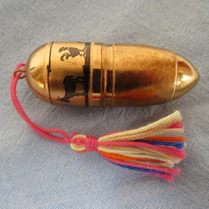 Puede incluir: Un recipiente de metal dorado con forma de bala que presenta una silueta negra de un gallo y una cabra. Un cordón rosa está unido a un extremo, conectado a una borla multicolor con hilos de lana de color crema, naranja, azul y rosa.