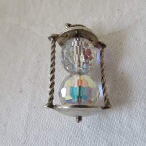 May include: A silver-toned charm in the shape of an hourglass. The charm features two faceted, iridescent crystal spheres stacked within a twisted metal frame. The top has a decorative flourish, and the bottom has a small, dangling element.
