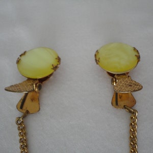 May include: A pair of gold-tone collar clips with a chain connecting them. Each clip features a round, pale yellow, translucent stone.