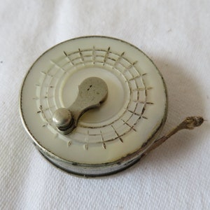 Op de afbeelding: Een vintage zilveren en witte meetlint met een decoratief ontwerp. De meetlint is rond en heeft een klein handvat.