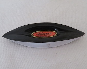 May include: A black tatting shuttle with a pointed, boat-shaped design. Centered on the top is an oval-shaped label with a red background and gold border, featuring the text Milward and the words MADE IN ENGLAND printed in black lettering.
