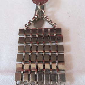 Puede incluir: Un portacadena de reloj de bolsillo de plata con una parte superior decorativa y una cadena hecha de cuadrados enlazados.