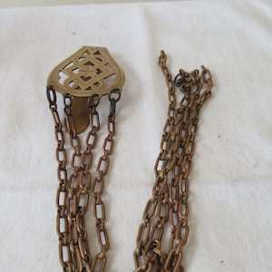 Op de afbeelding: Een gouden metalen ketting met een decoratieve, sierlijke hanger. De hanger heeft een geometrisch ontwerp in de vorm van een traan.