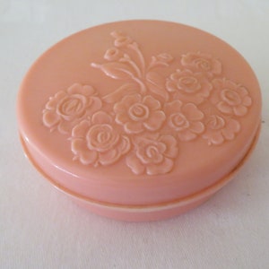 Può includere: Una scatola rotonda in plastica rosa con un design floreale in rilievo sul coperchio. Il design presenta un gruppo di rose e foglie.