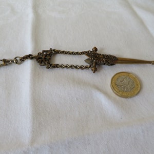 May include: A gold-toned pocket watch chain with a decorative sword-shaped fob. The fob is detailed with intricate floral designs and has a small chain attached to the top.