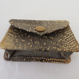 以下が含まれることがあります： ボタン留めの小さな茶色のヘビ革財布。財布は質感のある素材で作られており、前面に折り畳むフラップが付いています。