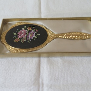 Könnte beinhalten: Ein goldfarbener Handspiegel mit einem ovalen Glas und einem floralen Design auf der Rückseite. Der Spiegel befindet sich in einer weißen Schachtel mit goldener Umrandung.