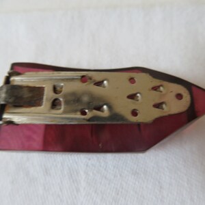 May include: A vintage purple glass brooch with a metal clasp. The clasp has a decorative design with small holes and a textured surface.