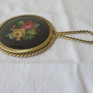 Könnte beinhalten: Ein kleiner, runder Handspiegel mit goldenem Rahmen, schwarzem Hintergrund und einem floralen Stickmuster. Der Spiegel hat einen gedrehten goldenen Griff.