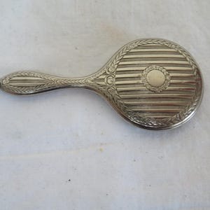 May include: A silver hand mirror with a decorative handle and a circular mirror with a raised design. The mirror has a striped pattern and a circular design in the center.