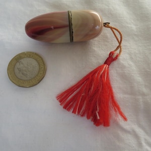 Puede incluir: Un pequeño recipiente ovalado con tapa abisagrada, con un patrón veteado en tonos rojos, rosas y crema. Una borla roja y blanca está unida. Una moneda está al lado. El recipiente mide unos 6 cm.
