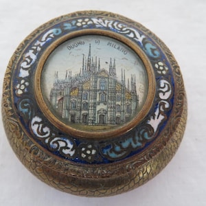 Puede incluir: Una caja de baratijas redonda de color dorado con un diseño de esmalte azul y blanco. La tapa presenta una pequeña imagen redonda del Duomo de Milán, una famosa catedral de Milán, Italia. El texto "DUOMO DI MILANO" está impreso alrededor de la imagen.