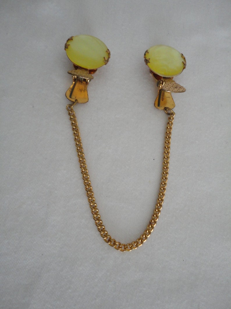 May include: A pair of gold tone clip-on earrings with a pale yellow oval shaped stone. The earrings are connected by a gold chain.