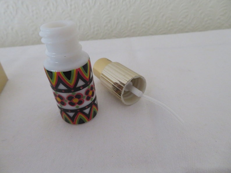 May include: A small, white glass bottle with a decorative floral and geometric pattern in red, green, yellow, and black. The bottle has a gold-colored cap and a white plastic tube. The cap is separate from the bottle.