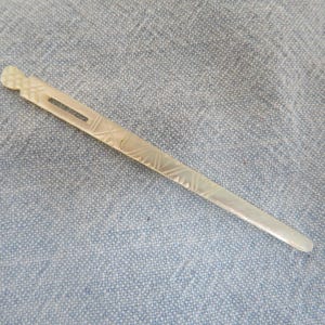Pode incluir: Um fino palito de cabelo esbranquiçado com uma cabeça decorativa esculpida e um recorte retangular. O palito tem padrões lineares gravados ao longo do seu comprimento. O acessório de cabelo é exibido em uma superfície de tecido azul.