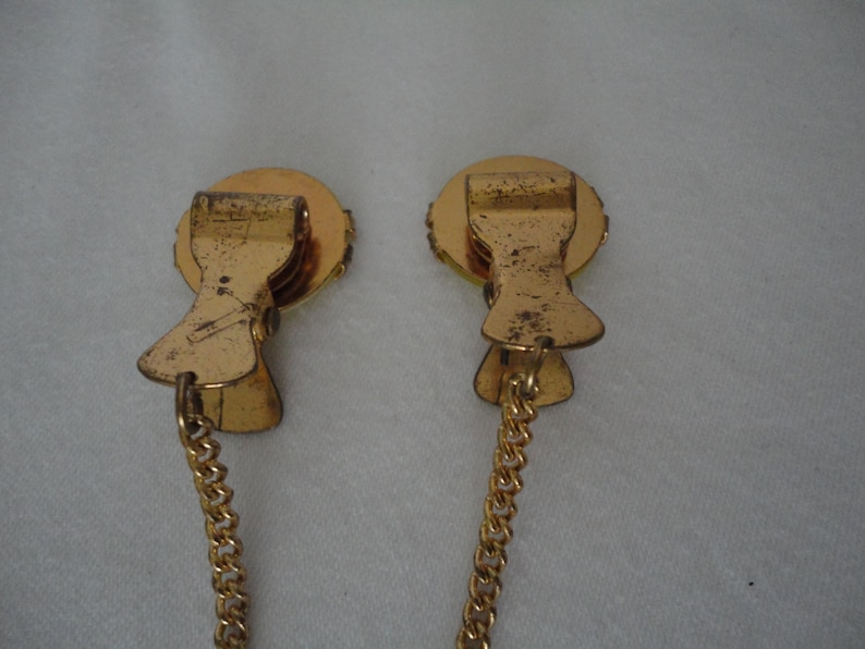 May include: A pair of gold-tone metal clips with a chain connecting them. Each clip has a round, decorative top with a raised design.