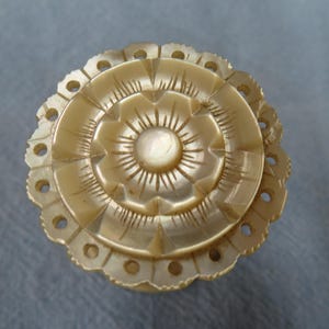 May include: A detailed, antique, ivory-colored trinket box lid. The lid features a central floral design with radiating lines and a scalloped edge with small circular cutouts. The overall design is symmetrical and ornate.